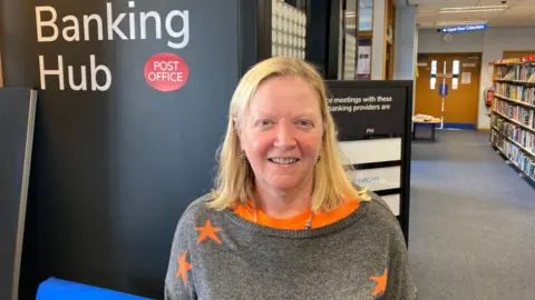 A woman wearing a grey and orange jumper with stars on it stands in front of a building and shelves of books