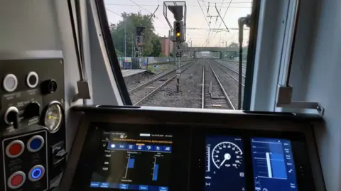 An inside of a train cabin, with a screen, and railway lines outside the cabin, with tracks, signals and a platform to the left. 