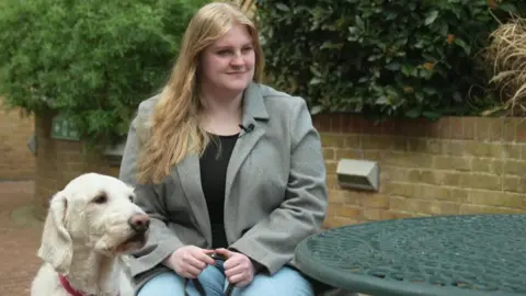 Jessica is sitting outside in a grey coat beside her cream-coloured guide dog