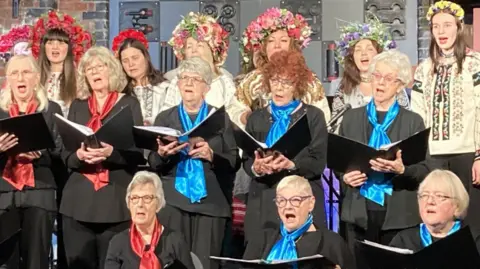 English and Ukrainian performers at the Ukrainian Christmas Service. Ukrainian singers are dressed in their national traditional costume during rehearsals. They have big floral head pieces, and white or light coloured shirt-style tops with embroidered detailing. The English singers are dressed in black shirts and trousers with red or blue silk neck ties. They are all holding song books