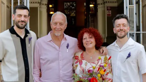 Four people standing outside a town hall for a wedding. Three males and one female. The woman is wearing a flowery dress and holding flowers. One three men are all wearing shirts with a purple flower attached to each. 