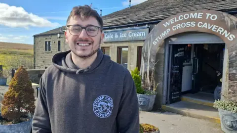 BBC/Seb Cheer A man, Oliver, smiles at the camera. He is standing in front of a building with a sign reading "Welcome to Stump Cross Caverns". Behind the building is a luscious green hill with blue sky above.