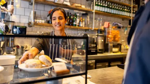 Getty Images Stock photo shows a cafe worker behind the till serving a customer who is partially out of view in front of the till, she is reaching for a plate of buns on display with coffee machines and orange press in the background amongst shelves full with stock.