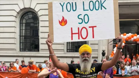 Manny Singh Kang He is holding a banner saying you look so hot and looking at the camera, while two runners in purple tops are also on the road behind him. Crowds of people are behind barriers in the background in front of a building.