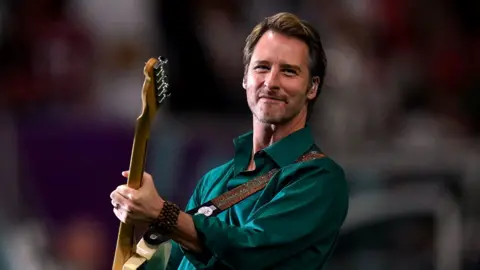 Chesney Hawkes performing with a guitar round his neck. He is wearing a dark green satin shirt and is smiling towards the camera.