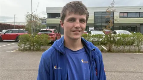 A young man wearing a blue T-shirt and hoodie with short brown hair stands in a car park with shrubs behind him. There is also a corrugated metal building behind him.