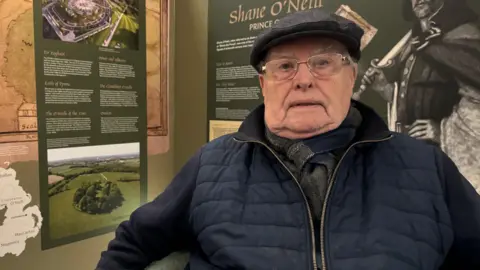 An older man is sitting in a corner looking into the camera. He is wearing metal rim glasses, a dark cap and scarf, and a navy coat. Behind him are information panels on the history of Shane O'Neill.