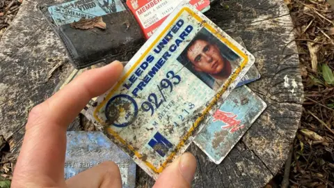The Secret Garden Rawdon A hand holding a Leeds United membership card which is covered in mud, there are some more cards and a wallet behind it balanced on a tree stump. 