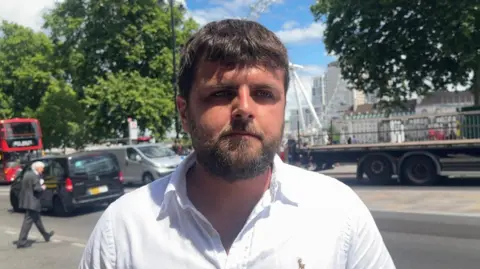 Electrician Sonny Spencer is wearing a white polo shirt and a beard. There is traffic behind him