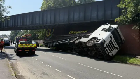 A lorry wedged under a blue railway bridge, with its trailer partially crushed. Highways officers are parked nearby.
