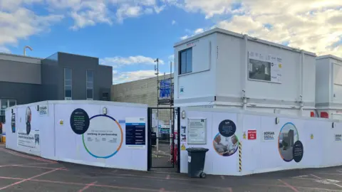 Amy Holmes/BBC A construction area outside Milton Keynes hospital is enclosed by white hoarding covered with partnership signage and safety notices, with stacked portable site offices and a clear blue sky above.