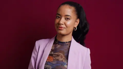 A woman stands against a red backdrop. She is wearing a pink suit jacket and a multi-coloured top. She has straight black hair.