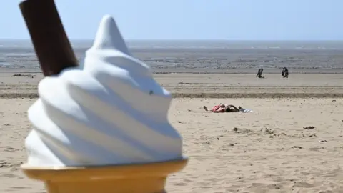 Getty Images Close up image of an ice cream cone, topped with a flake. Behind, and more in focus is a beach scene, with a sunbather on the sand and four people walking by the shore, with the sea in the background.