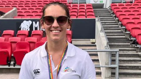 Tania smiles at the camera from behind large dark sunglasses. She has brown hair pulled back in a ponytail, and wears a rainbow lanyard and a white polo shirt with the City Academy logo on it.