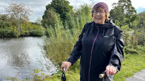 Julia Bryson/BBC Sheila Mahon dressed in walking clothes with sticks, next to Ilkley Tarn.