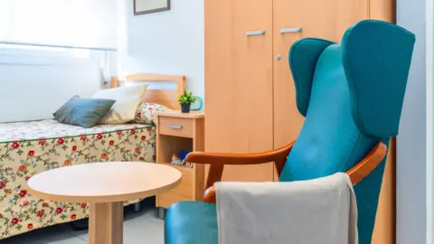 A small care home room consisting of a single bed with floral covers, a blue arm chair and light brown wardrobe and bedside table. On top of the bedside table sits a small potted plant. 