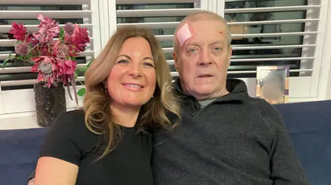 Kirsty Jewson is smiling and has long brown hair with blonde highlights and a black top. Brian Jobson is sitting next to her and has short grey hair, a green top and grey jumper. He has a plaster on his head. They are sitting on a blue sofa. There is a window behind them and white horizontal blinds with a dark glass vase with red flowers and a photo frame on the white windowsill.