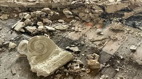 A plaster scroll lies on the floor next to bits of rubble that has fallen from a water damaged wall and ceiling.