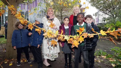 Mayor of Sunderland Allison Chisnall cuts the ribbon to open Mowbray Park Community Garden. She is standing with councillor Beth jones and five school children. The ribbon has been decorated with autumnal leaves.