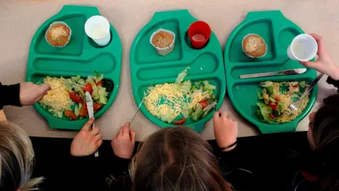 Children eating school dinners on a table. Lettuce leaves, tomatoes and cheese is visible.  A cup with squash is on the tray along with a pudding item. 