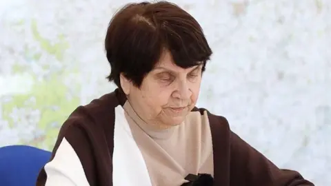 Norfolk Police Galina sitting on a blue chair is looking down reading a statement. She has short brown hair and is wearing a beige, white and brown jumper.