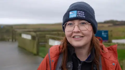 A woman wearing a dark blue beanie hat and orange waterproof jacket stands on a wooden boardwalk at a nature reserve. Binoculars hang around her neck. Behind her are grassy fields, a wooden fence, and a winding path under an overcast sky.