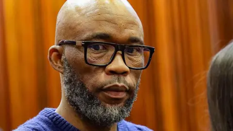 A head and shoulders picture of Cat Matlala in court - he is wearing a blue jumper, the neckline of which can just be seen at the bottom of the picture.