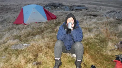 Amanda Higgins Amanda is sitting on the grass next to a pitched tent. She is sipping from a mug.