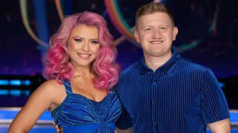 Getty Images Sam Aston and Molly Lanaghan smiles for the camera on the Dancing on Ice set. Aston is wearing a blue-and-black striped shirt and he has ginger hair and blue eyes. Lanaghan is wearing a sparky blue dress and has pink hair.