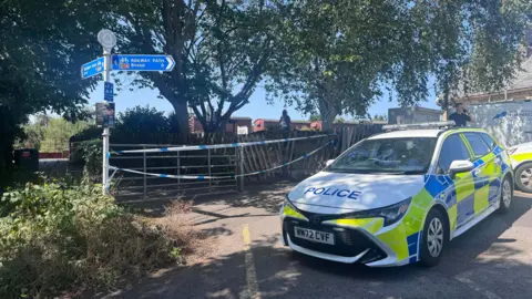 A police car parked up near a gate leading to the Bristol and Bath Railway Path. There is blue and white police tape across the gate.