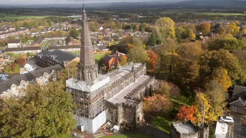 Historic England Aerial drone view of church with spire above scaffolding surrounding the main base of the building. Autumnal trees and a graveyard are outside the church with houses and countryside around.