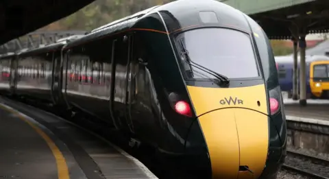 A train with 'GWR' on the front pulling into a platform with red lights.