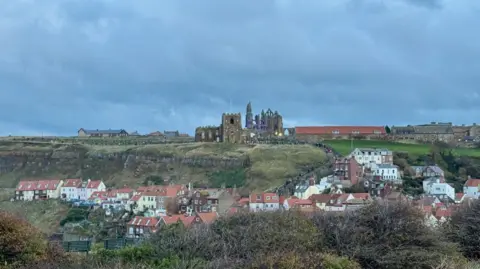 A ruined abbey in the distance behind a church. Low rise buildings near the abbey, which is on a grassy clifftop. Buildings in the valley beneath the clifftop.
