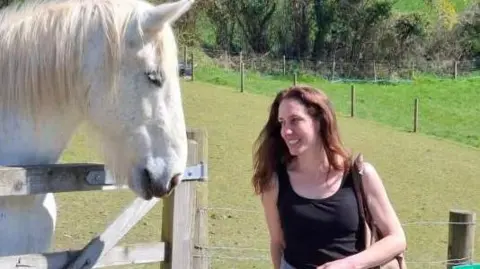 Belinda Taylor is stood wearing a black sleeveless vest with long brown hair. She is stood by a wooden gate with a white horse on the other side. 