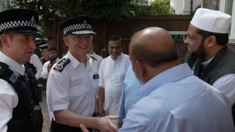 Met Police Commissioner Mark Rowley - wearing a white shirt and police hat - shakes hands with a man in Muslim dress. They stand among a group of people including another police officer and worshippers.