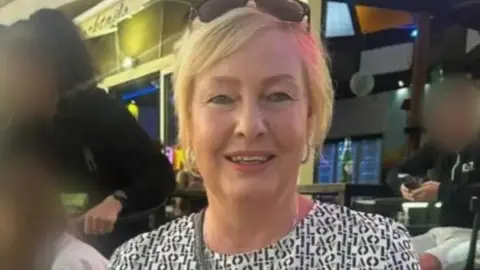 Yvonne Ford smiling at the camera with sunglasses on her head and blonde hair, pictured in a public place with people's faces blurred out in the background