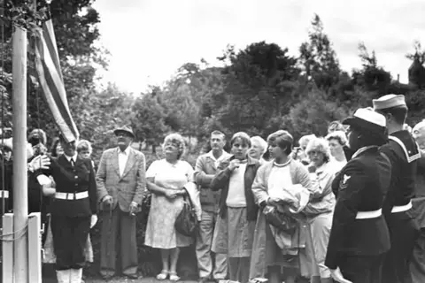 National Trust A black and white image of a dozen people standing around a flag pole as an American flag is raised. Some people are looking up at it, while others are looking around. There are people in uniform who are saluting as the flag as it goes up. There are tall, lush trees behind them.