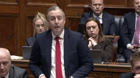 NI Assembly A man in a navy suite and pink tie, has short grey/brown hair. Half a dozen men and woman sit behind him on blue-leather, wooden benches. 
