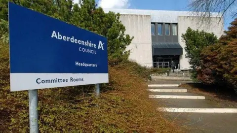 Aberdeenshire Council's Woodhill Headquarters. The building is behind a set of stairs and green and brown trees and bushes. A blue and white sign which says "Aberdeenshire Council Headquarters Committee Rooms" is at the front of the picture. 