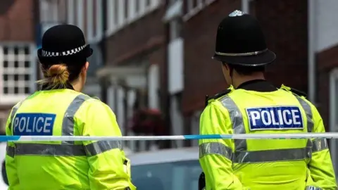 Two police officers, a woman on the left and man on the right, stand on the other side of blue and white police tape stretched across the area behind them. In front of them is the blurred outline of a building.