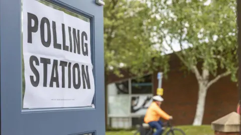 A polling station poster on the glass of an open door.- it reads "Polling Station" in black font on a white background. In the background there is a cyclist that is out of focus. They are wearing an orange coat and blue trousers and are cycling past two trees and a green lawn.