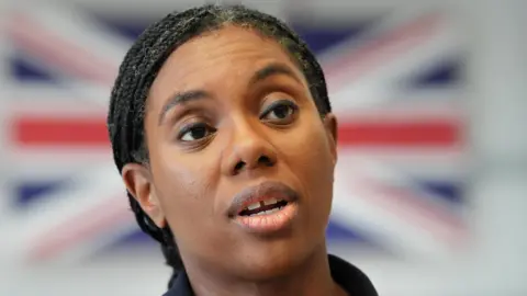Kemi Badenoch close up with a blurred UK flag behind her