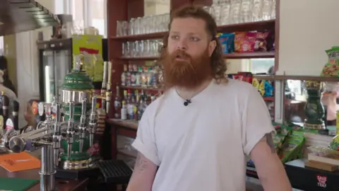 Man with long hair and a large beard standing behind a pub bar next to several beer taps, with shelves of glasses and snacks in the background.