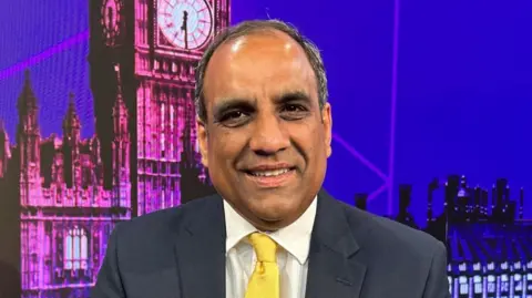 Shaffaq Mohammed, leader of the Liberal Democrats in Sheffield, sits in front of a digital backdrop showing Big Ben in London. He is smiling, wearing a dark suit, and yellow tie.