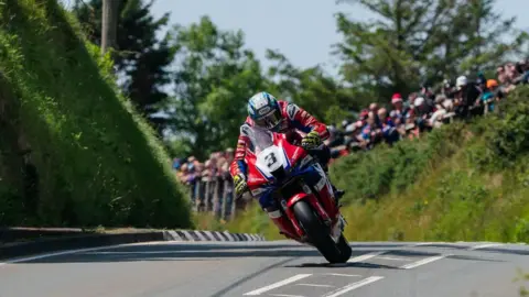 ISLE OF MAN TT A rider on a red bike with the number three on it, it tilts over as it takes a corner, there are people lining bushes.