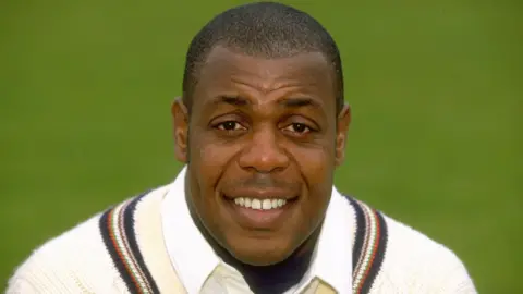 David 'Syd' Lawrence. He is has brown eyes and short dark hair. He is pictured wearing a white cricket jumper with a stripe around the neck. He is looking at the camera and smiling. 