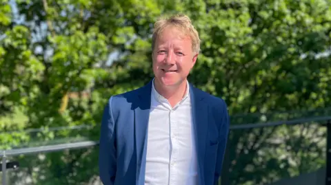 Head and shoulders image of Paul Bristow. He has light hair, a blue suit jacket and white shirt and is standing in front of trees.