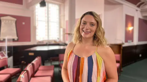 Laura Pick has long curly blonde hair and is smiling. She is wearing a colourful striped summer dress as she stands in the Methodist Centre.