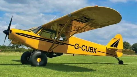 A picture of an aircraft, a G-CUBX, issued by the Air Accidents Investigation Branch. The yellow plane is standing on grass with trees behind it and it is a sunny day.