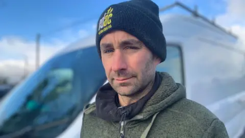 A man who is wearing a black beanie hat is looking at the camera. He's wearing a green fleece with a black snood around his neck. He is standing in front of a white van.